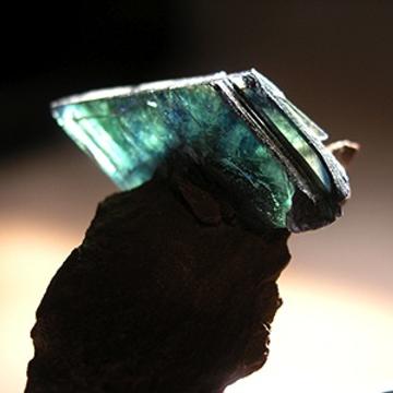 a transparent, blue-green crystal of vivianite; back-lit to better display colouring. The mineral displays a darker blue about the centre, where it's attached to the matrix.