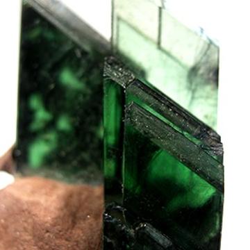 Translucent green crystals of vivianite atop a sandy matrix. There are three vertical crystals; one each in the foreground, mid-ground, and background, becoming increasingly out of focus.